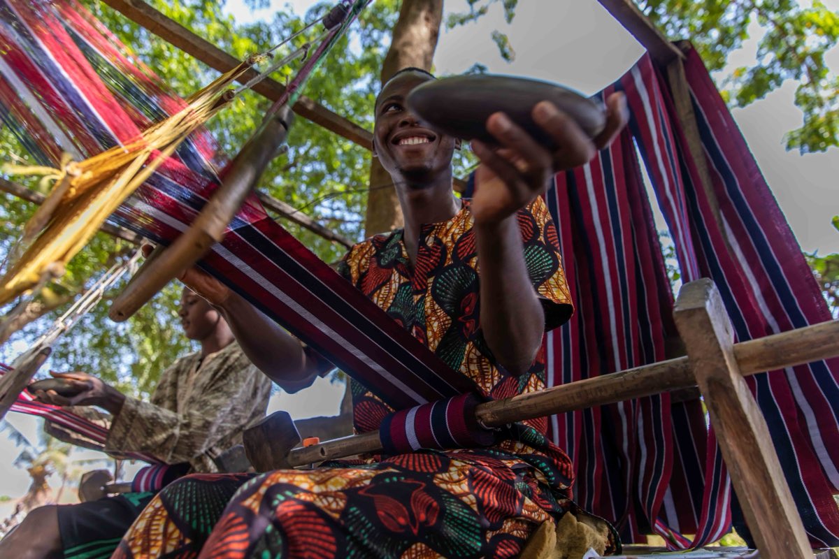 A Yoruba weaver at a narrow-strip loom, shuttle in hand, striped aso-òkè stretched on the frame around him.