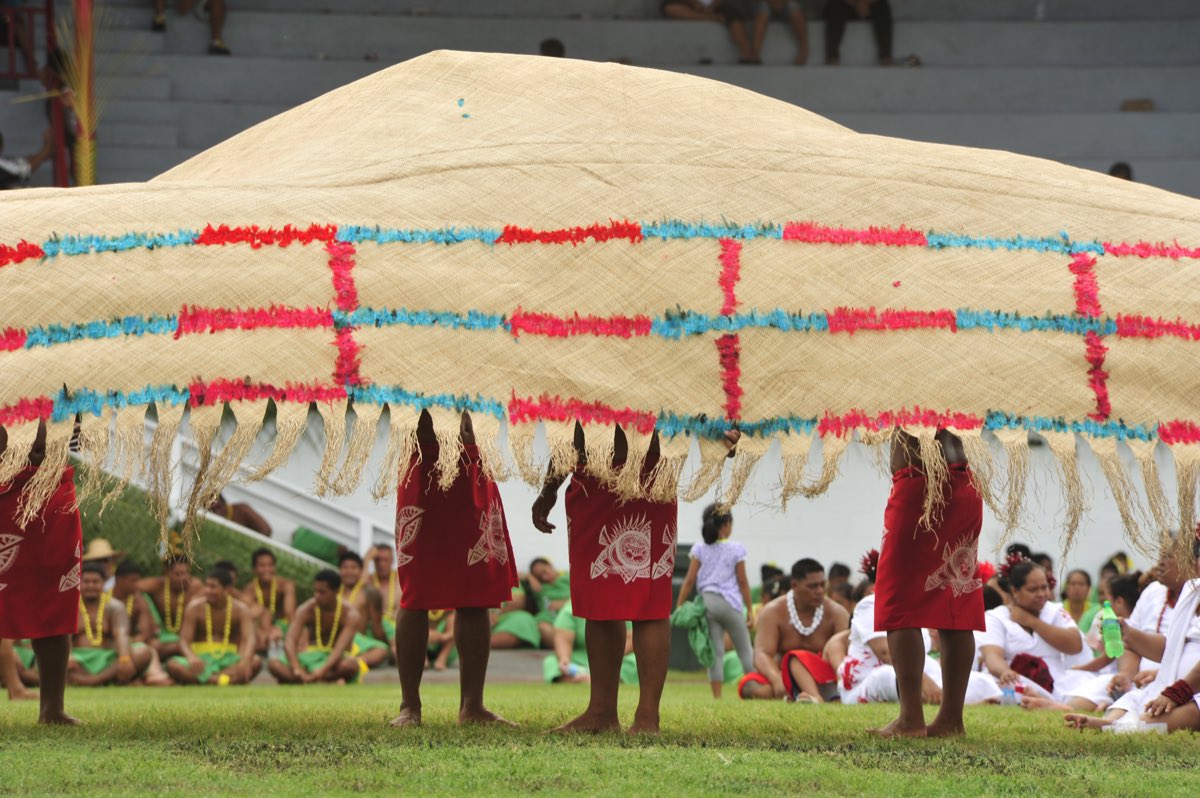 An enormous ʻie tōga fine mat, edged with red feathers, held aloft at a ceremony in the Samoan islands.