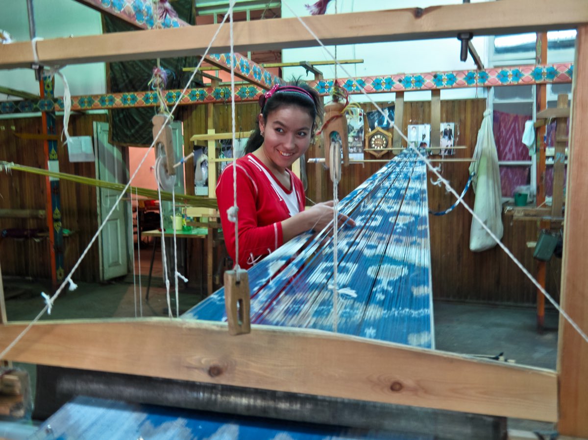 A weaver at her loom in Margilan, Uzbekistan, behind a long span of resist-dyed ikat warp threads.