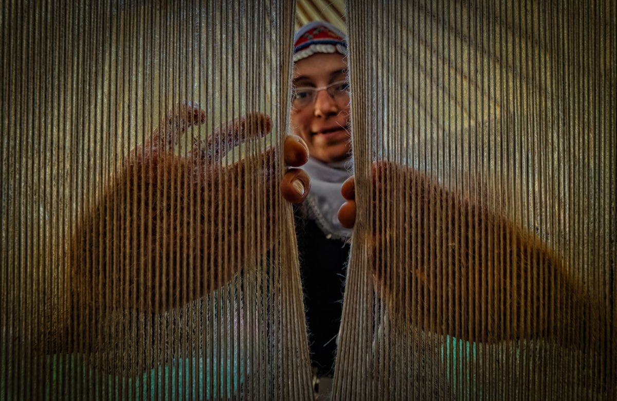 A Turkish weaver seen through the vertical warp threads of her kilim loom, hands reaching between the strings.