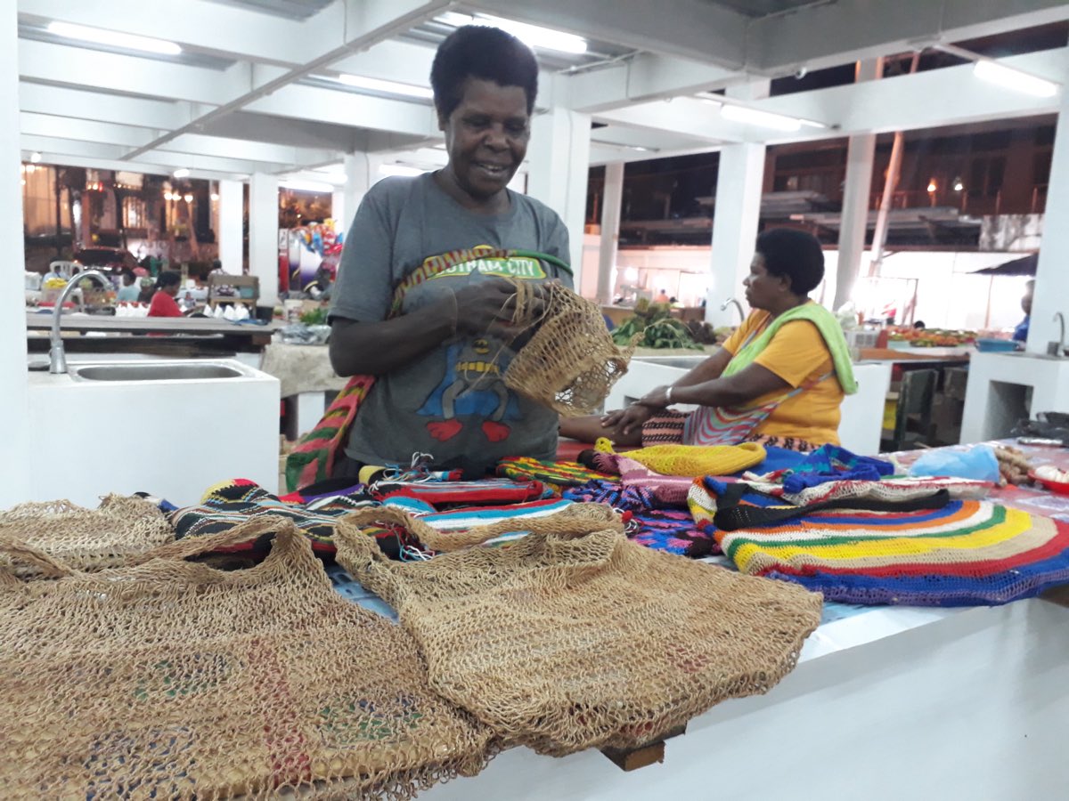 A Papuan weaver hand-knotting a noken at her market stall, finished net bags draped in the foreground.